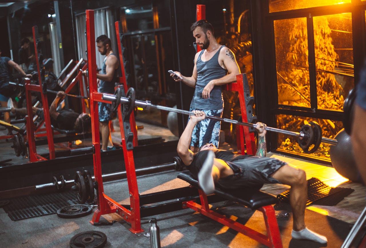 Man bench pressing in a modern gym while another checks smartphone. Fitness and technology theme.